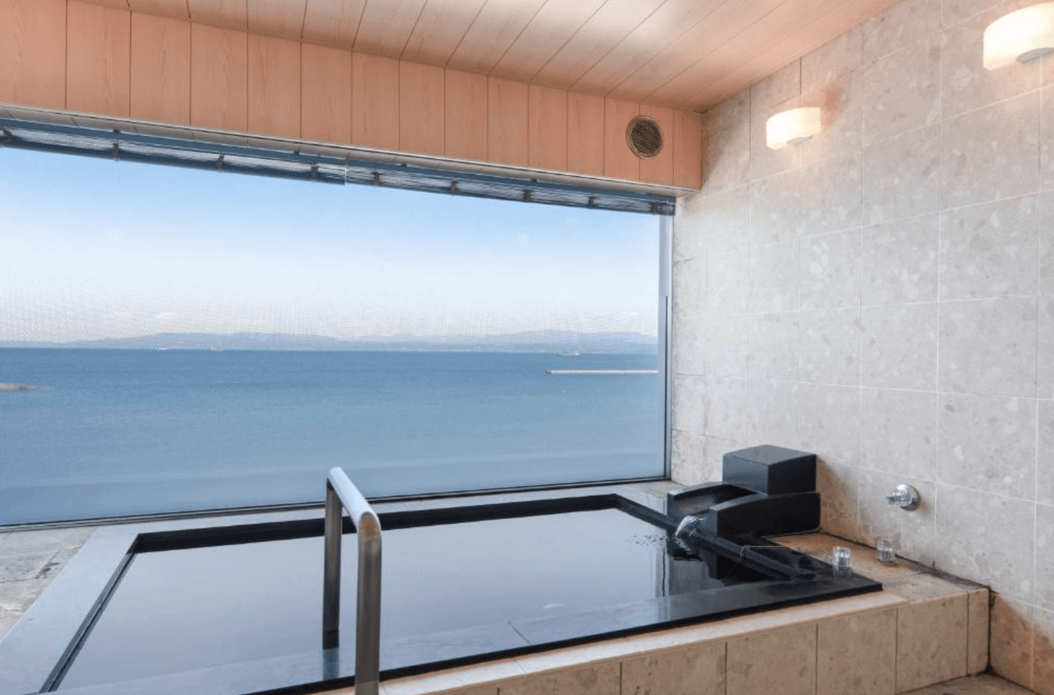 【指宿住宿】指宿海上飯店 (Ibusuki Kaijyo Hotel) 的景觀大浴場，設有黑色的方形石造浴池，旅客可透過寬闊的玻璃窗，一邊享受溫泉的療癒，一邊欣賞窗外平靜無波的海面風光。