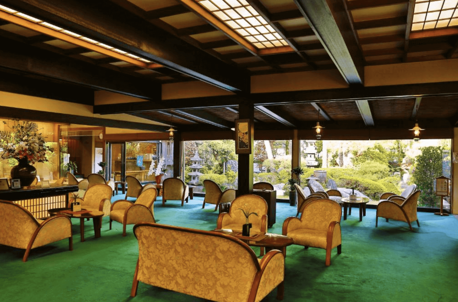 【指宿住宿】指宿秀水園旅館 (Ryokan Ibusuki Syusuien) 的大廳交誼廳，擁有挑高的深色木樑天花板與整面落地窗，窗外即是精心修剪的日式庭園美景，搭配復古優雅的黃色沙發椅，展現老牌旅館的氣派。
