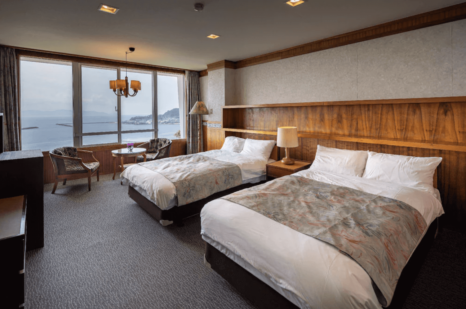 【指宿住宿】指宿鳳凰飯店 (Ibusuki Phoenix Hotel) 的海景雙床客房，設計典雅穩重，設有寬敞的木質床頭櫃與舒適雙人床，窗邊配置了休閒桌椅區，讓旅客能輕鬆坐著眺望錦江灣的遼闊海景。