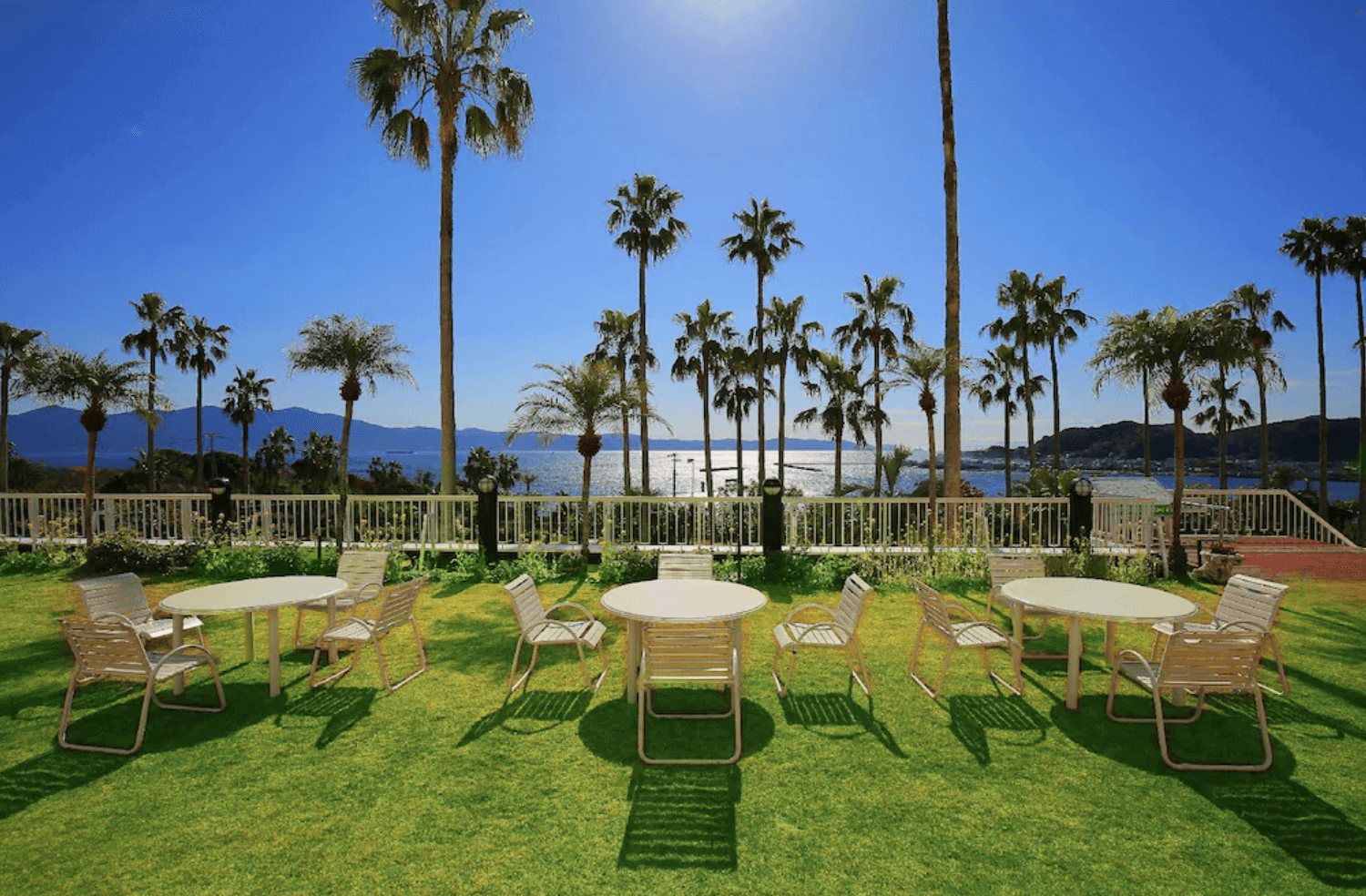 【指宿住宿】指宿鳳凰飯店 (Ibusuki Phoenix Hotel) 的戶外花園與景觀休息區，綠意盎然的草坪上擺設了白色戶外桌椅，四周環繞著高大的椰子樹，背景即是藍天與大海，視野極佳。