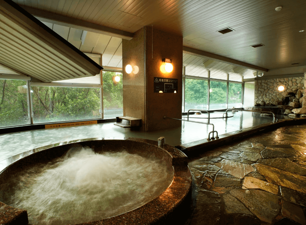日勝生加賀屋 (Radium Kagaya) 的大眾湯「望月」，擁有寬敞的石造溫泉浴池，搭配大面景觀窗引入戶外自然光與綠意，營造出寧靜且充滿禪意的泡湯環境。