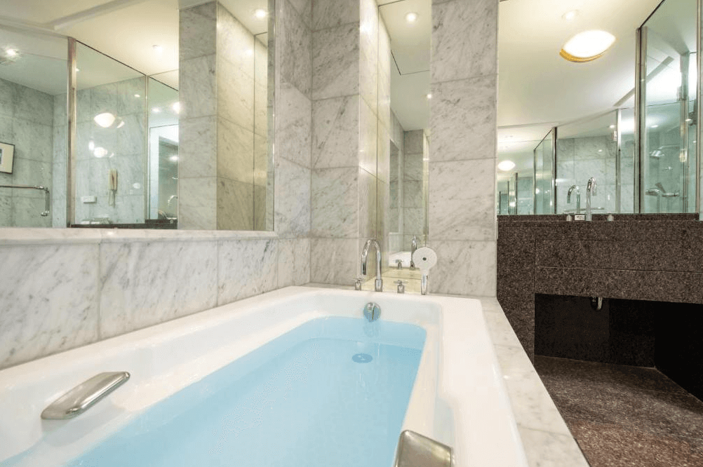 【大分住宿】日航大分綠洲塔酒店 (Hotel Nikko Oita Oasis Tower) 的大理石衛浴空間,牆面與地面皆採用淺色大理石鋪設,配備寬敞的浴缸與明亮的鏡面設計,展現經典星級飯店的豪華質感與潔淨空間。