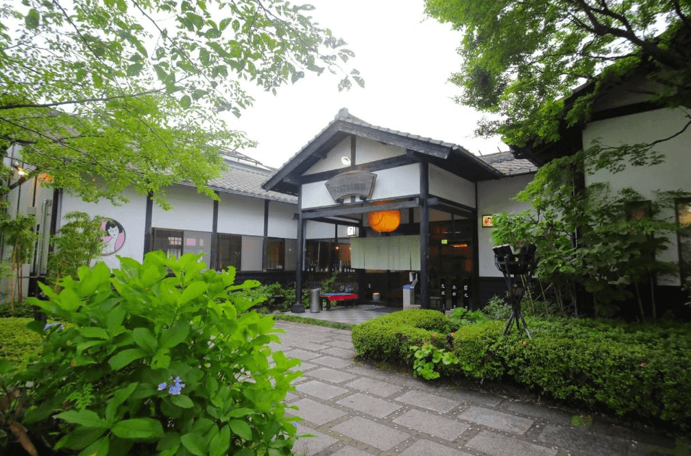 【霧島住宿】櫻櫻溫泉旅館的日式傳統外觀，入口處掛有暖簾與燈籠，建築周圍環繞著綠意盎然的樹木與石板步道，充滿濃厚的溫泉鄉風情。