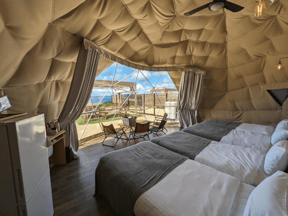 【沖繩南城住宿】沖繩八點度假村 (8POINT RESORT OKINAWA) 的豪華露營圓頂帳篷客房，內部空間寬敞，挑高的米色幾何帳篷頂下並排擺放著三張舒適床鋪，充滿溫馨的野奢露營氛圍。