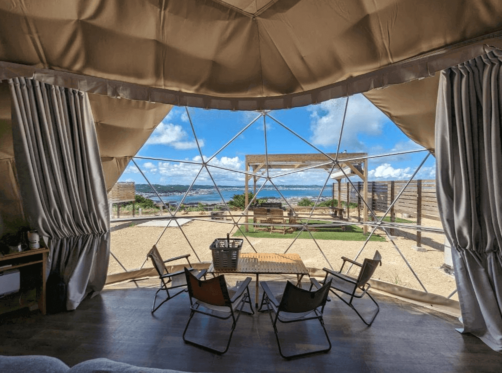 【沖繩南城住宿】沖繩八點度假村 (8POINT RESORT OKINAWA) 的圓頂帳篷休憩區，透過巨大的幾何透明景觀窗，旅客可坐在室內的露營椅上，愜意地眺望戶外露台與遠方湛藍的沖繩海景。
