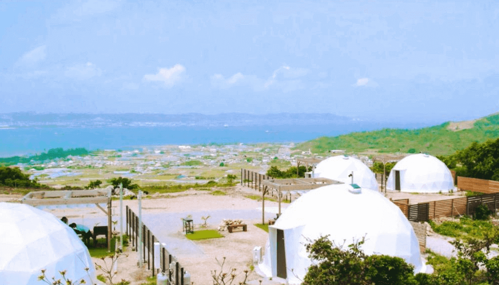【沖繩南城住宿】沖繩八點度假村 (8POINT RESORT OKINAWA) 的園區景觀，座落於綠意盎然的山丘之上，白色的幾何圓頂帳篷群與後方遼闊的藍天碧海相互輝映，展現絕佳的度假視野。