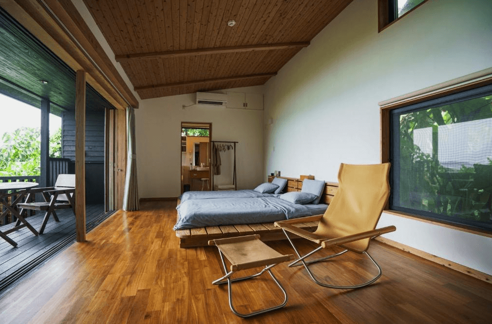 【沖繩南城住宿】海濱茶館旅館 (Villa SACHIIBARU) 的獨棟木屋客房，室內採用溫潤的天然木材裝潢，配備兩張低矮的舒適床鋪與設計感搖椅，落地窗外連接著綠意盎然的專屬木棧露臺。
