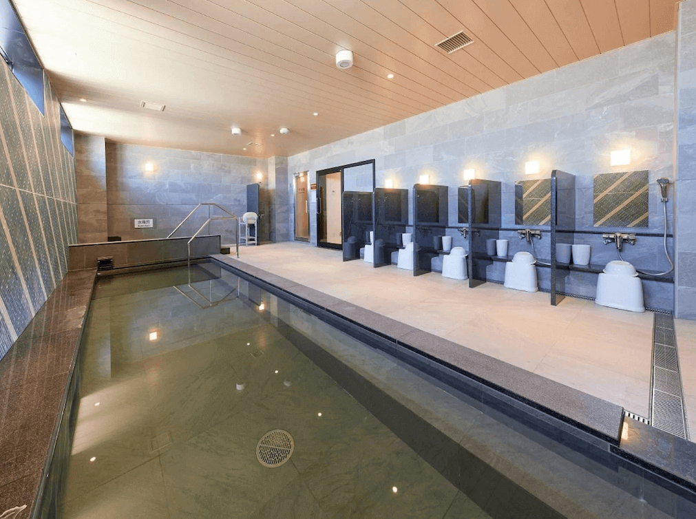 【鹿兒島住宿】海茵娜飯店鹿兒島天文館 (Henn na Hotel Kagoshima Tenmonkan) 的大浴場空間，採用沉穩的灰色石磚設計，設有長型寬敞的溫泉浴池與一整排獨立的淋浴沖洗區，環境乾淨舒適。