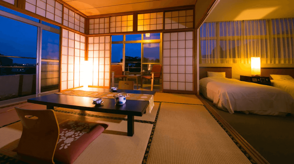 【屋久島住宿】「田代旅館分館」的和式房型，鋪有榻榻米，中央擺放傳統日式矮桌與座墊，大片落地窗外可見夜景，房間以柔和燈光營造溫暖氛圍。