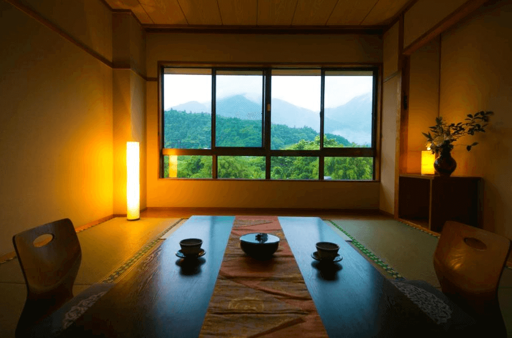 【屋久島住宿】「田代旅館分館」的和室空間，面向大片山景窗景，房內設有日式桌椅，柔黃燈光搭配綠意盎然的山景，呈現靜謐悠閒的住宿環境。