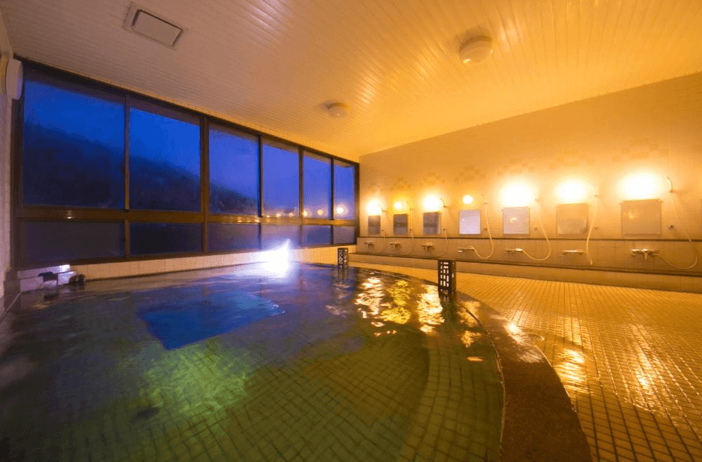 【屋久島住宿】「田代旅館分館」的大浴場，設有大面積景觀玻璃窗，可遠眺夜間山景，洗浴區整潔明亮，溫泉池水面反射柔和燈光，營造放鬆氛圍。