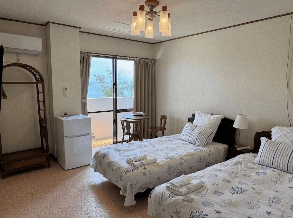【沖繩南城住宿】米巴魯海濱旅館 (Beachside Pension Mi-baru) 的西式雙床客房，鋪設印有貝殼圖案的床單呼應海濱主題，房內搭配復古的藤編衣櫃與古典吊燈，落地窗外即是可欣賞蔚藍海景的專屬陽台。