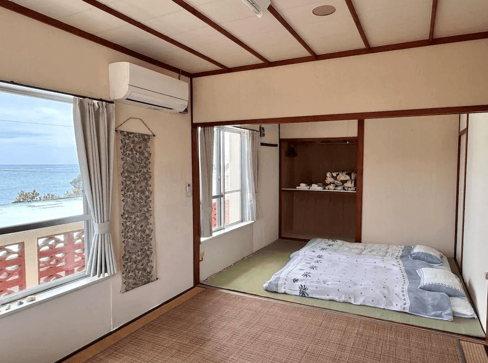 【沖繩南城住宿】米巴魯海濱旅館 (Beachside Pension Mi-baru) 的簡約和室客房，在榻榻米上鋪設了日式床墊，空間樸實溫馨，透過寬敞的窗戶，旅客躺在房內即可眺望戶外平靜的海洋景致。