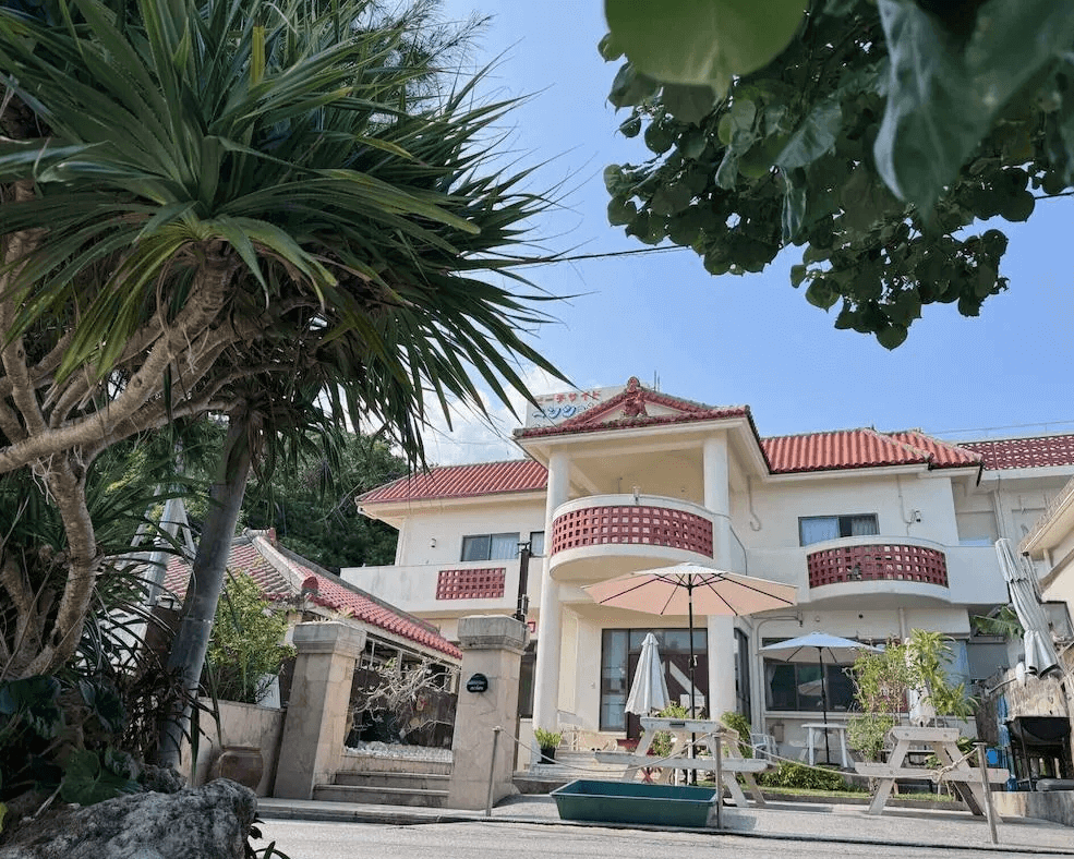 【沖繩南城住宿】米巴魯海濱旅館 (Beachside Pension Mi-baru) 的外觀建築，為典型的沖繩風格白牆紅瓦屋頂設計，庭院中種植著茂盛的熱帶林投樹並設有戶外野餐桌椅，洋溢著悠閒自在的南國民宿風情。