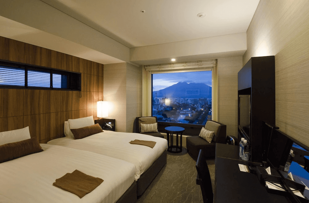 索拉利亞西鐵飯店鹿兒島 (Solaria Nishitetsu Hotel Kagoshima) 的精緻雙床客房，房內運用深色木紋飾板營造沉穩氛圍，透過大面觀景窗，旅客可在房內欣賞夜晚燈火通明的鹿兒島市景與櫻島火山輪廓。