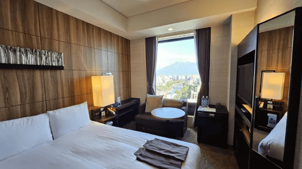 【鹿兒島住宿】索拉利亞西鐵飯店鹿兒島 (Solaria Nishitetsu Hotel Kagoshima) 的舒適客房，床頭牆面飾有獨特的藝術裝置，窗邊設有休憩沙發區，窗外明亮的日光映照著流經市區的甲突川與遠方壯麗的櫻島火山。