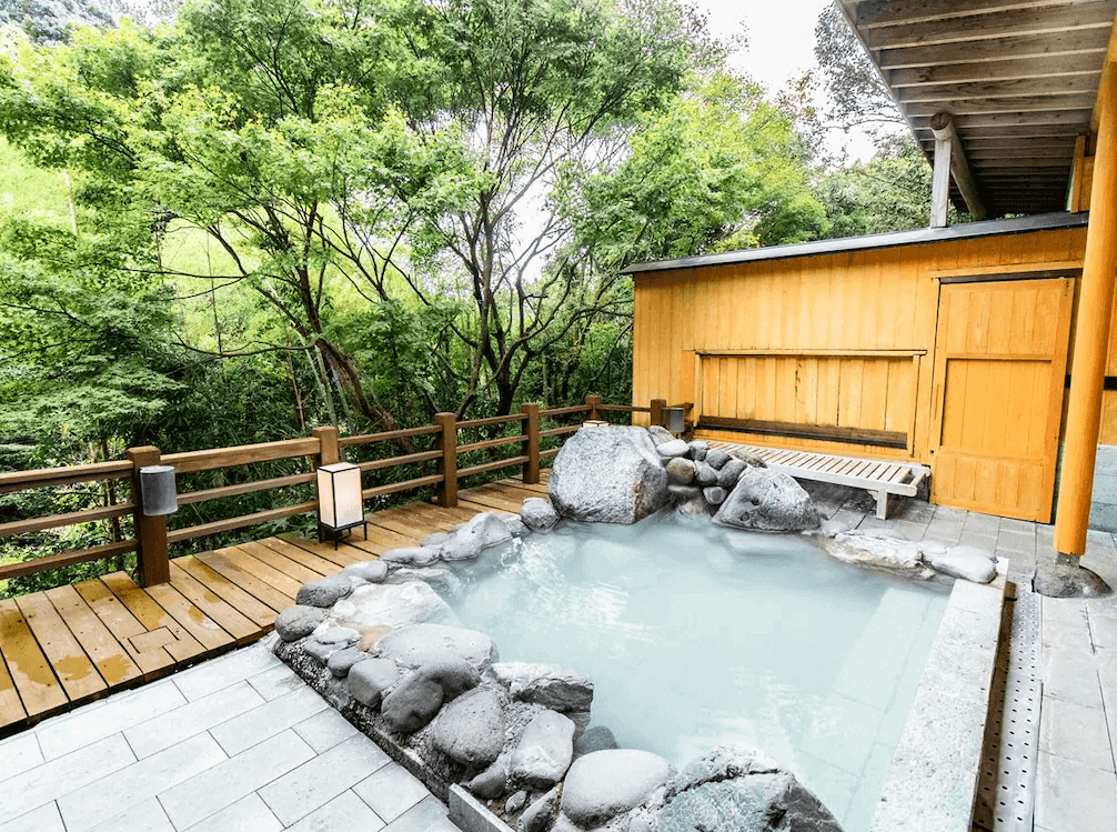 【霧島住宿】霧島紅葉谷清流莊的露天風呂，以天然岩石砌成的溫泉池坐落於木棧平台上，四周被茂密的樹林環繞，讓旅客能在大自然中盡情放鬆。