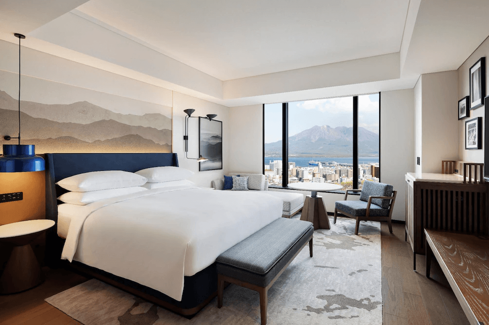 【鹿兒島住宿】鹿兒島喜來登 (Sheraton Kagoshima) 的景觀雙人客房，床頭飾有山水意象畫作並搭配藍色吊燈，房內靠窗處設有休閒桌椅區，讓旅客在房內即可正面眺望櫻島火山與錦江灣的絕美景致。