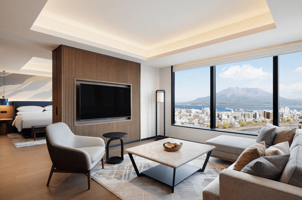 【鹿兒島住宿】鹿兒島喜來登 (Sheraton Kagoshima) 寬敞的豪華套房客廳，配備淺灰色L型沙發與大理石茶几，木質格柵電視牆區隔出臥室空間，透過整面落地窗可飽覽櫻島火山與鹿兒島市區的壯闊全景。