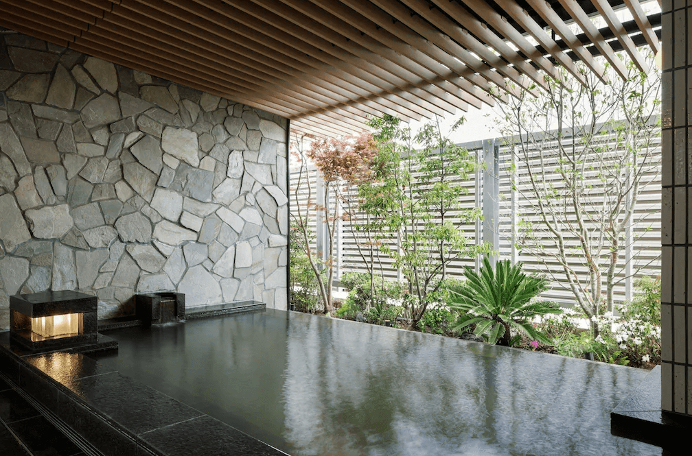 【鹿兒島住宿】鹿兒島喜來登 (Sheraton Kagoshima) 的日式溫泉浴場，以天然石材砌成的牆面搭配黑色石造浴池，半露天的設計結合木質格柵與庭園綠意，提供旅客寧靜放鬆的泡湯環境。