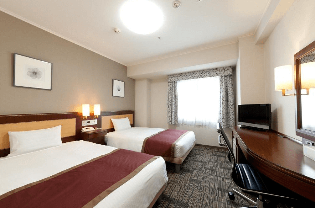 【鹿兒島住宿】鹿兒島法華俱樂部飯店 (Hotel Hokke Club Kagoshima) 的明亮雙床客房，配備兩張單人床與沿牆設計的長條形辦公桌區，簡約典雅的裝潢風格提供旅客機能完善的休憩環境。