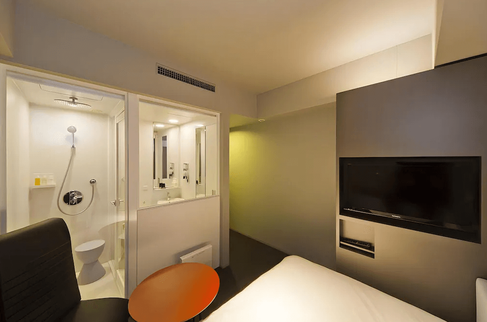 【鹿兒島住宿】鹿兒島雷姆飯店 (Remm Kagoshima) 的現代客房視角，特色為透明玻璃隔間的衛浴設計，配有舒壓淋浴花灑（Rain Shower），房內亦配置了按摩椅與壁掛式電視，展現機能性十足的空間利用。