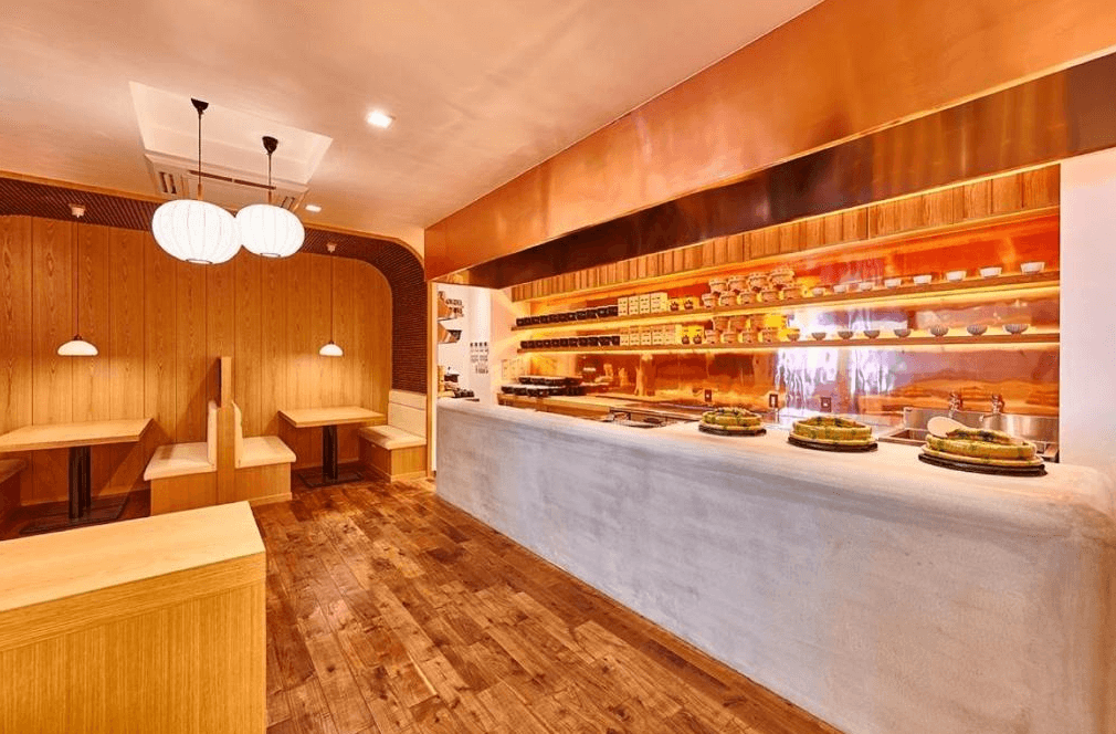 【鹿兒島住宿】鹿兒島雷姆飯店 (Remm Kagoshima) 的餐飲吧台區，採用大量的溫暖木質裝潢搭配柔和的球形吊燈，水泥質感的取餐檯上擺放著精緻的料理，營造出溫馨愜意的用餐氛圍。