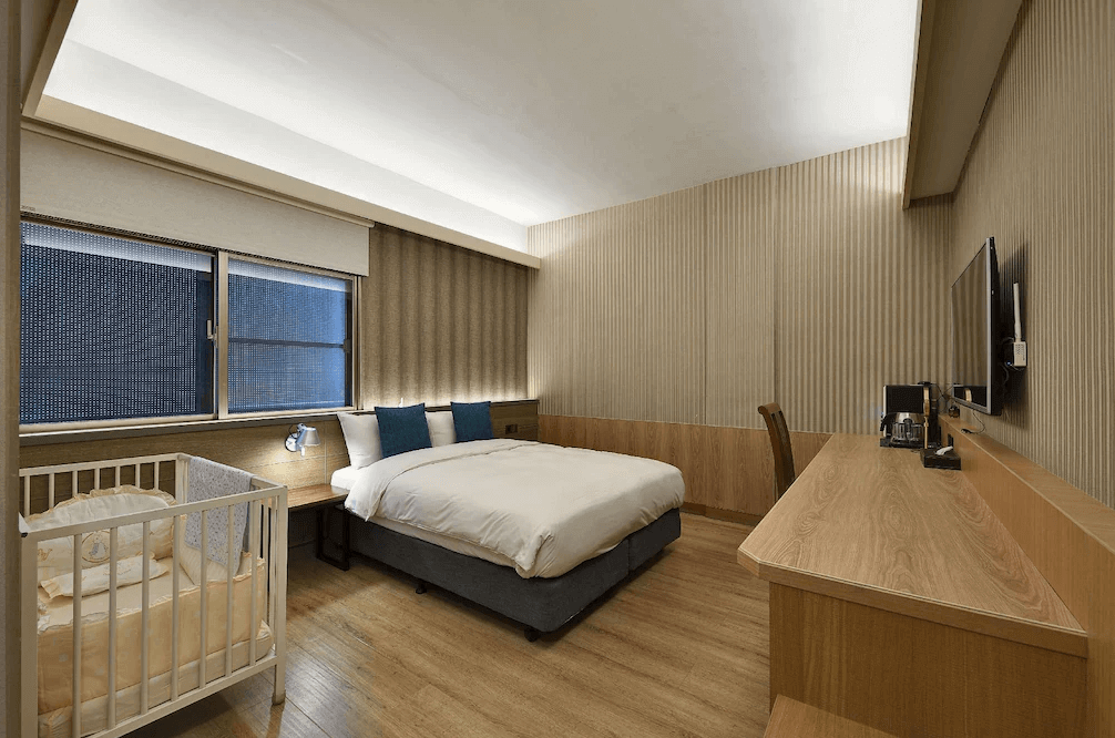 EG Hostel溫馨親子房，木質調空間設計典雅舒適，配置一張雙人床與長型木作書桌，最貼心的是房內備有專屬嬰兒床，非常適合新手爸媽帶著寶寶入住。