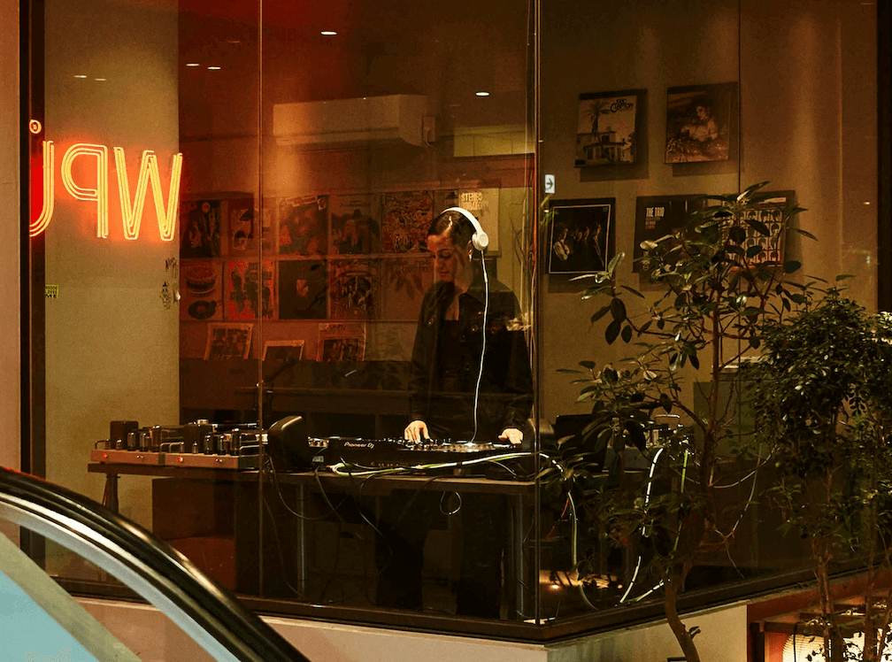 WPÜ飯店新宿的夜晚DJ音樂展演空間,透過玻璃櫥窗可見DJ正在播放音樂,牆面展示黑膠唱片並搭配醒目的紅色霓虹燈標誌,展現結合音樂與住宿的獨特生活風格。