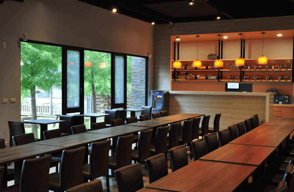 一峸一宿民宿景觀餐廳，用餐區採光極佳，透過大面窗戶可欣賞窗外種植的落羽松與綠地，配置多張深色長桌供多人用餐，自助吧台區設計簡約明亮。