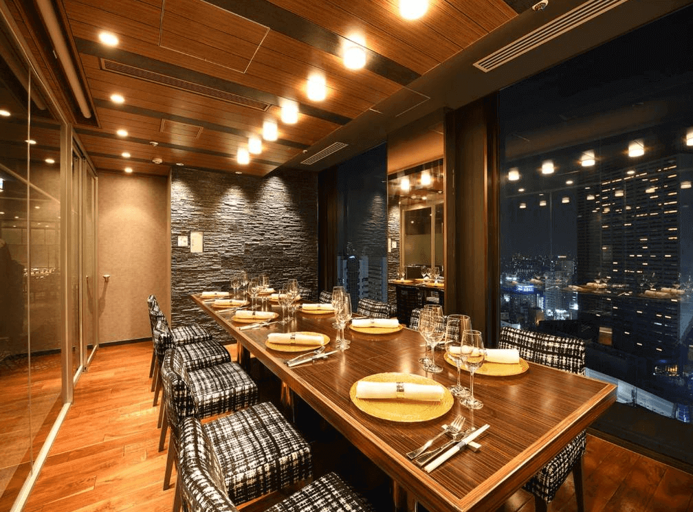 新宿王子大飯店的高樓層景觀餐廳包廂,長桌鋪設整齊餐具,搭配千鳥格紋餐椅與深色石材牆面,窗外即是璀璨迷人的新宿夜景,用餐氛圍私密且高級。