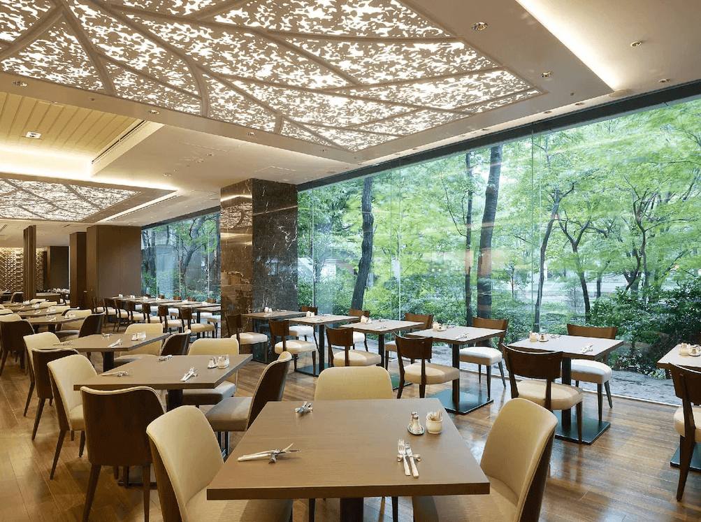 東京京王廣場飯店的景觀餐廳,採用挑高天花板與大片落地玻璃窗設計,讓窗外蓊鬱的森林綠意與室內樹影圖騰天花板融為一體,用餐環境明亮且充滿生機。