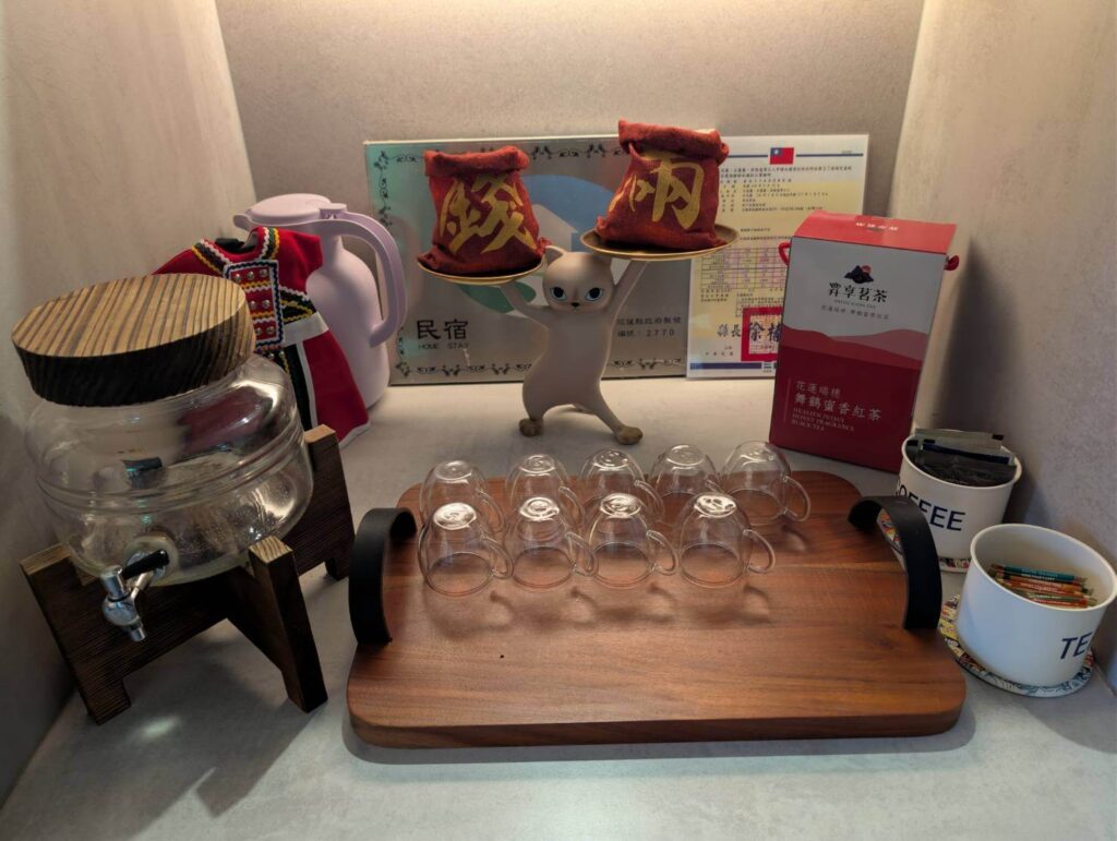 自然野奢1樓備品：溫馨的茶水備品區，桌面上擺放花蓮瑞穗舞鶴蜜香紅茶禮盒、精緻雙層玻璃杯組與復古玻璃飲水罐，後方陳列民宿登記證與可愛招財貓擺飾，提供住客在地風味茶飲與咖啡包。