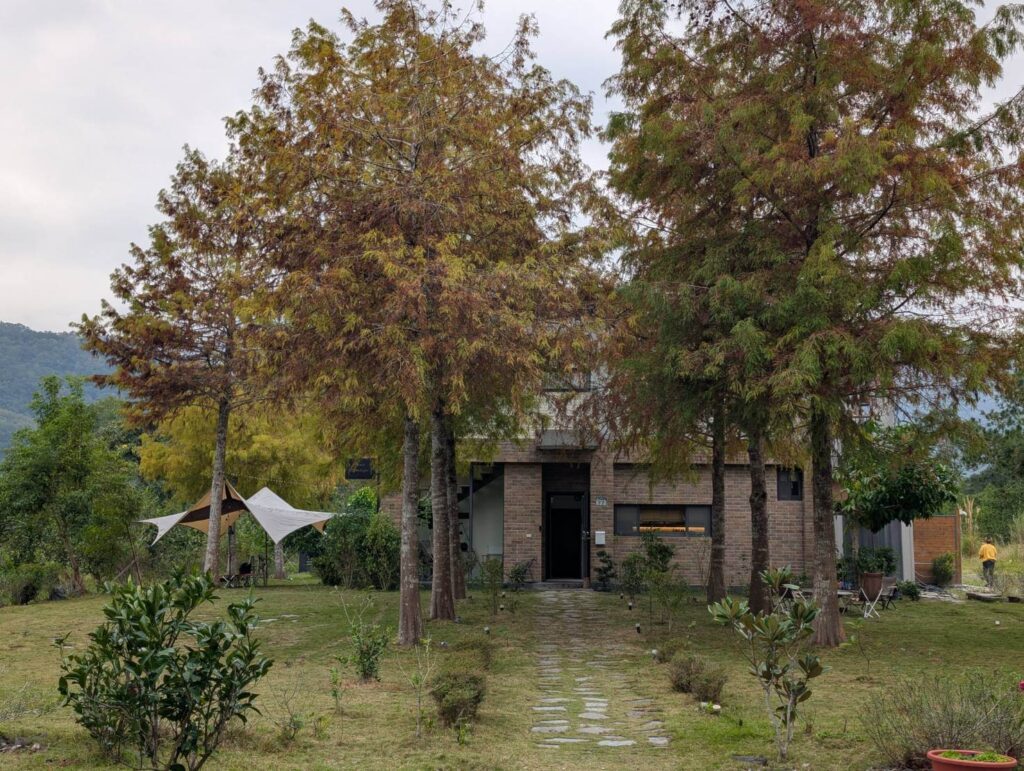 "自然野奢精選示意圖"自然野奢戶外環境，紅磚民宿建築隱身於轉紅的高聳落羽松林間，前方擁有寬敞草地與石板步道，左側搭建白色戶外天幕帳篷，背景群山環繞，呈現靜謐悠閒的度假氛圍。