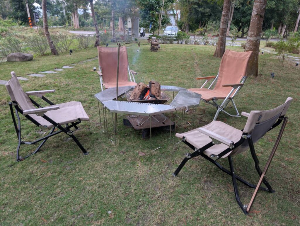 自然野奢篝火：戶外草地上設置專業不鏽鋼六角焚火台與圍桌，周圍環繞四張舒適的卡其色露營椅，讓住客能在林蔭間安全體驗生火樂趣，享受圍爐取暖的療癒時光。