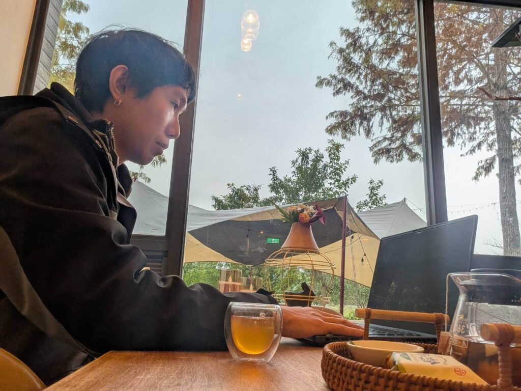 自然野奢長桌辦公：生活口帶部落格創作者，於大面落地窗前的原木長桌使用筆電工作，窗外綠意盎然的樹林與露營帳篷景致盡收眼底，桌旁擺放溫熱茶飲與點心，展現結合工作與度假的愜意氛圍。