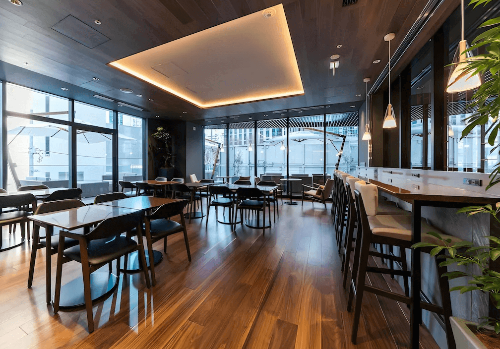 西新宿大和羅伊內特高級飯店的公共交誼廳與用餐區,擁有溫暖的木質天花板與照明,大面落地窗連接戶外露台,室內亦設有配備插座的長桌區,非常適合商務辦公。