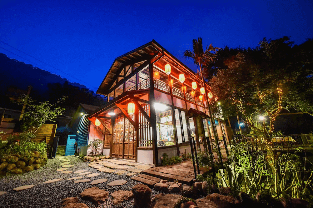 【苗栗寵物友善住宿】響水客棧 (Xiang Shui Inn) 的迷人夜景外觀，這棟兩層樓的復古木造建築在藍調夜色下顯得格外溫馨，屋簷下懸掛著一整排紅燈籠，搭配大面玻璃窗透出的暖黃燈光與戶外石板庭園，營造出宛如電影《神隱少女》般的懷舊氛圍