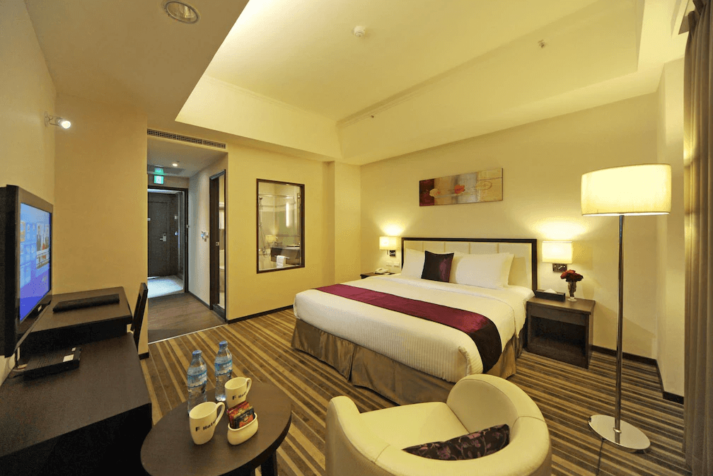 F Hotel 花蓮站前館的精緻雙人房實景,房內配有一張寬敞的加大雙人床、簡約木質書桌及暖色調落地燈,條紋地毯營造出溫馨的住宿氛圍。