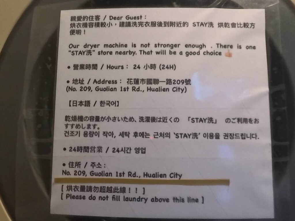 小旅行迷你公寓的多國語言公告單。公告內容主要提醒住客因公寓內的烘衣機容積較小，建議洗完衣服後可前往附近的「STAY洗」自助洗衣店烘衣。單據上列出了該店的營業時間為 24 小時，以及詳細地址：花蓮市國聯一路209號。公告內容包含了繁體中文、英文、日文及韓文四種語言，最下方則有醒目的「烘衣量請勿超越此線！！」警告標語。