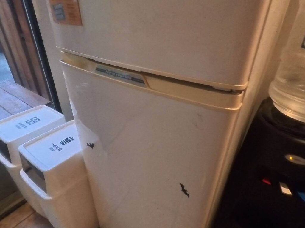 小旅行迷你公寓的白色雙門小冰箱。冰箱頂部貼有一張橘色小標籤，中段把手處印有「Joy in your life」的字樣，機身下層門板上有幾個深色的標記。冰箱左側鄰近落地窗處，放置了兩個標有「一般垃圾」等字樣的白色分類垃圾桶；右側則緊鄰一台飲水機，上方放有大桶裝水。整體呈現出簡潔、實用的居家或公寓住宿空間配置。