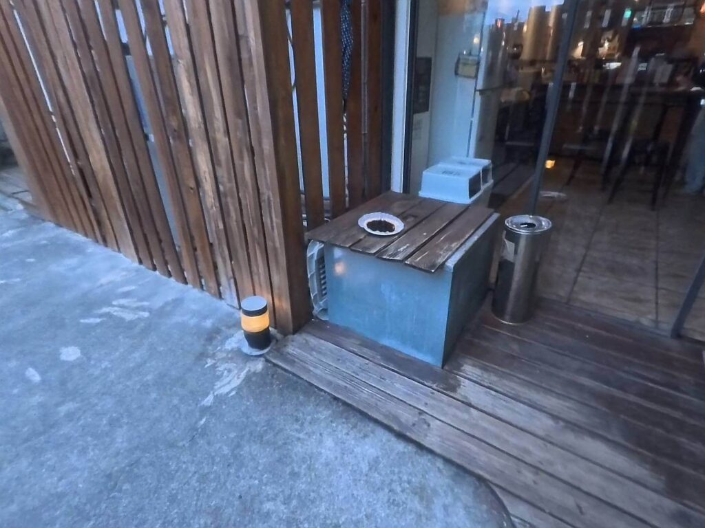 小旅行迷你公寓的室外吸菸區，鄰近玻璃門入口，設有不鏽鋼菸灰缸與木質遮蓋的基礎設施，環境簡潔明亮。