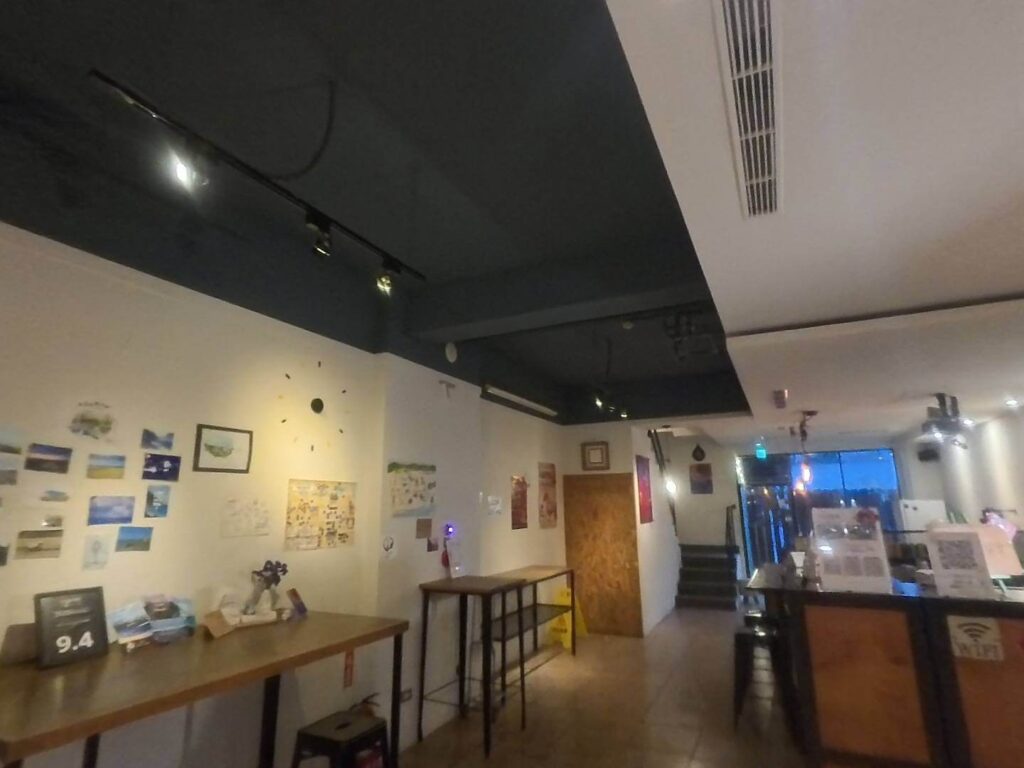 小旅行迷你公寓大廳空間：室內裝潢採現代簡約工業風，天花板為深色調搭配軌道燈照明。牆面佈置了多張風景明信片、地圖與裝飾畫，並展示著 9.4 分的高評價牌卡。空間內設有木質高腳桌椅與櫃檯，營造出溫馨且具設計感的旅宿氛圍。