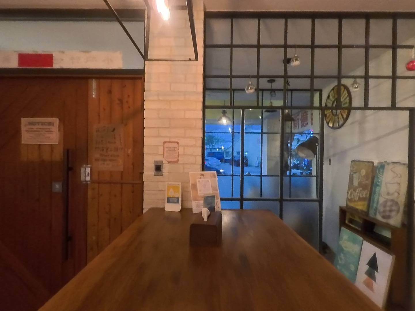 小旅行迷你公寓大大桌空間：室內採用溫潤的木質長桌，背景結合工業風格的黑色格狀落地窗與白磚牆設計。空間中裝飾著復古咖啡標誌與文藝掛畫，搭配柔和的暖色燈光，營造出寧靜且適合放鬆或交流的質感氛圍。
