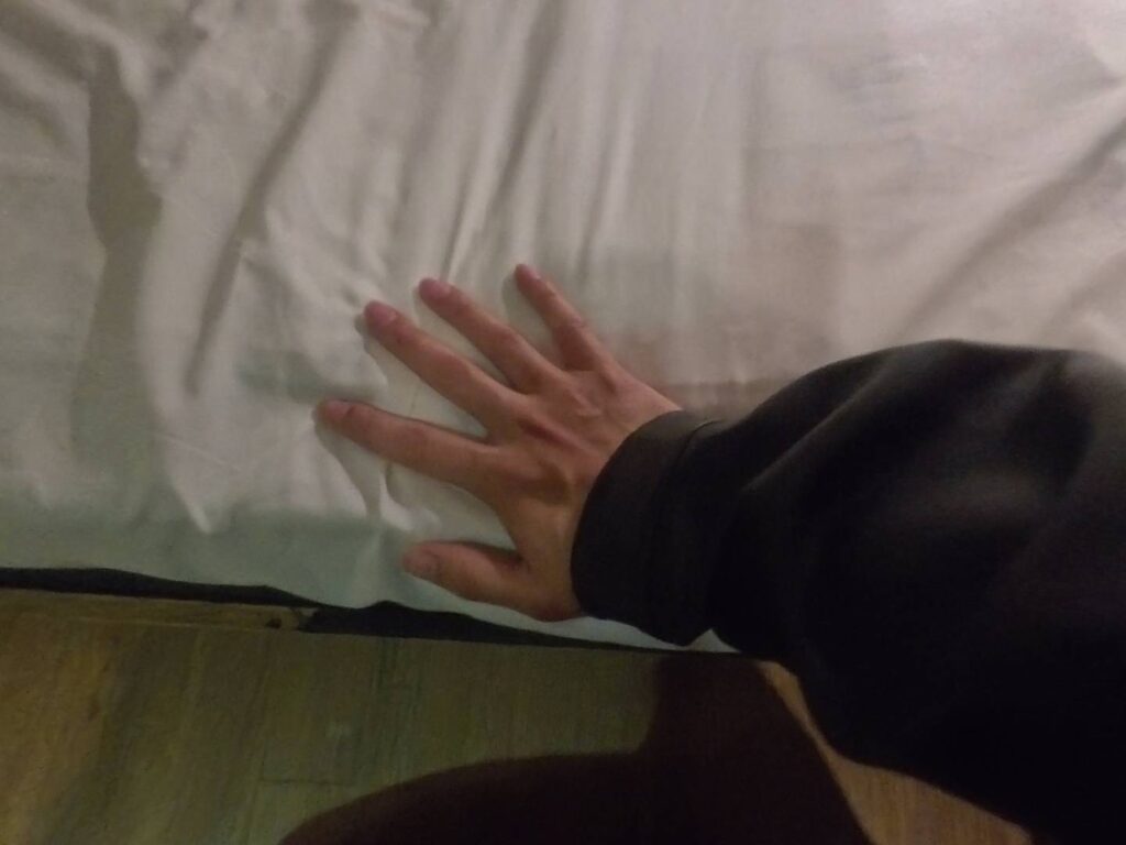 小旅行迷你公寓的床墊細節。畫面中一隻手正在按壓鋪著白色床單的床墊，以此展示床墊的軟硬度與彈性。