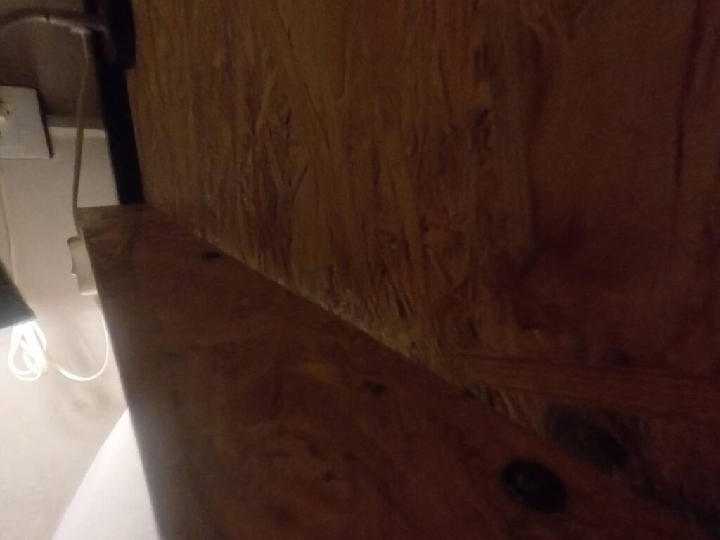 小旅行迷你公寓的床頭置物架。特寫展示了採用粗獷風格木紋板材（OSB 定向纖維板）製成的床頭邊架，旁邊可見電源插座。