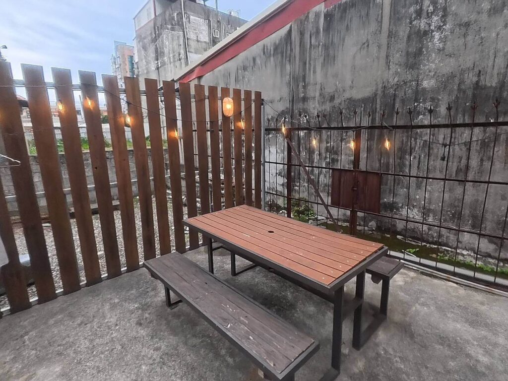 小旅行迷你公寓的戶外露台區，設有木質長桌與長凳，背景為木柵欄裝飾，上方掛有暖色調的工業風串燈，營造出溫馨放鬆的氛圍。
