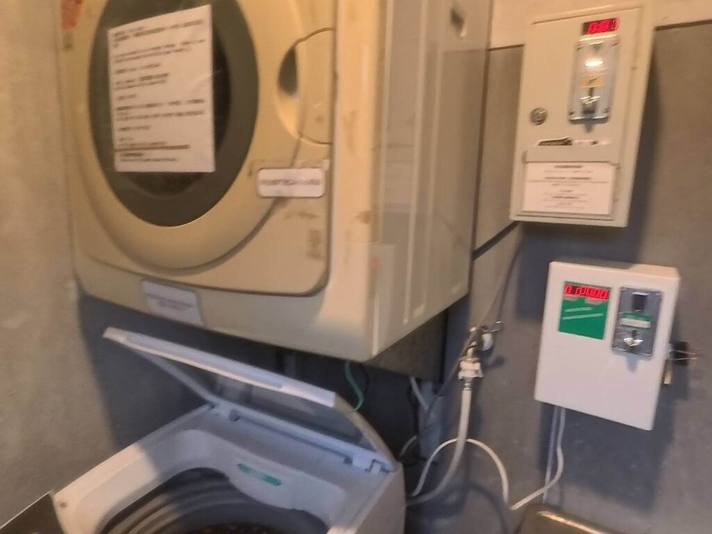 小旅行迷你公寓的投幣式洗烘衣設備。畫面下方是一台開啟上蓋的直立式洗衣機，上方則懸掛著一台圓形透明窗門的烘衣機，機器上貼有使用說明告示。牆面上安裝了兩個投幣控制箱，上方箱體設有紅色數字顯示器，顯示目前狀態或餘額。整體環境呈現簡潔的工業風格，牆面為灰色調，是典型公寓或青年旅館提供的自助洗滌空間。