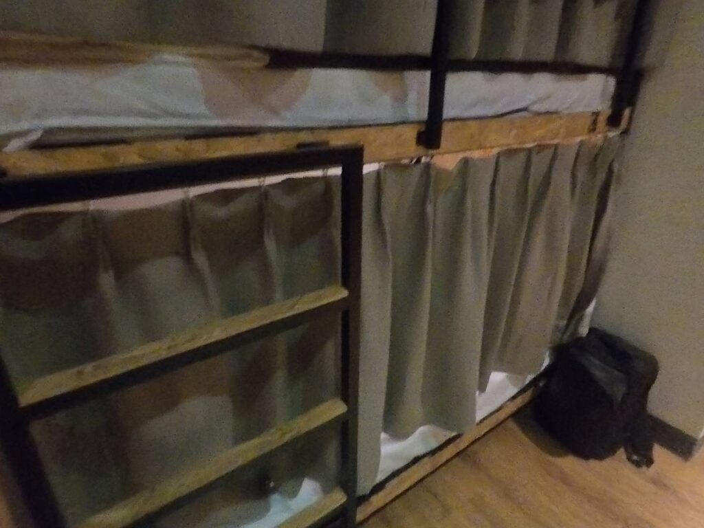「小旅行迷你公寓（Mini Voyage Hostel）的背包客房內部，呈現木質雙層床位，下層床鋪設有遮光簾以維護隱私，床邊配有黑色金屬爬梯。」