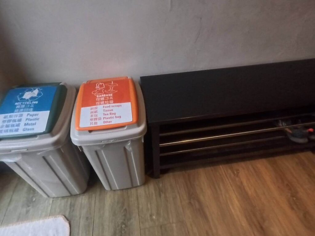 「小旅行迷你公寓的一角，靠牆擺放著一藍一橘蓋子的資源回收與垃圾桶，旁邊則是一個黑色的多層開放式鞋架，地板為淺色木紋材質。」